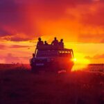 a vibrant sunset illuminates a diverse group of travelers atop an open safari vehicle, surrounded by the breathtaking expanse of tanzania's savanna teeming with wildlife.